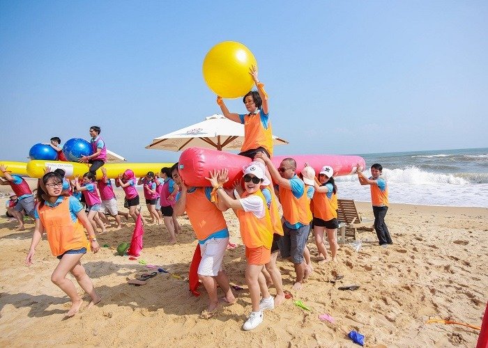 Nhóm người chơi team building trên bãi biển với áo cam và xanh, đang tương tác cùng các quả bóng màu sắc.