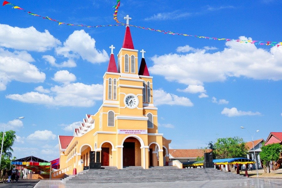 Cao Nguyên Vân Hòa: Khám Phá 'Đà Lạt Thứ 2' Của Xứ Nẫu Với Những Điều Đặc Biệt 3 Nhà thờ gạch đỏ nổi bật với nhiều tháp chuông, đồng hồ lớn, nắng sáng và bầu trời trong xanh trang trí bằng cờ dây.