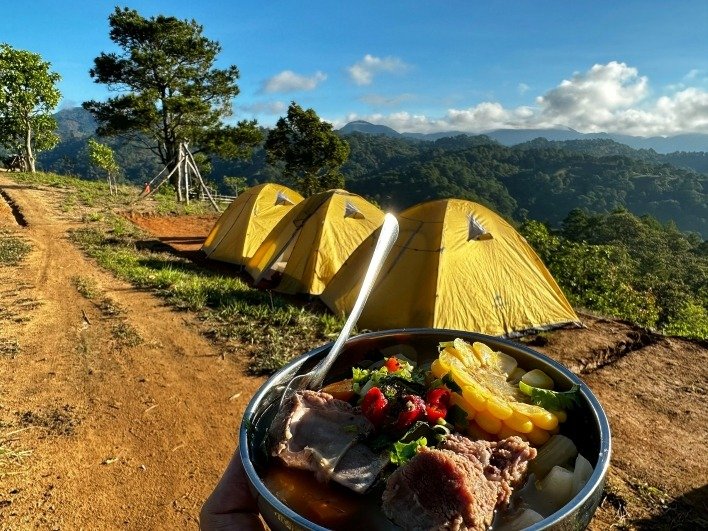 Bữa ăn cắm trại ngon lành trước cảnh núi non tuyệt đẹp với lều trại vàng dưới bầu trời xanh, tạo cảm giác yên bình.