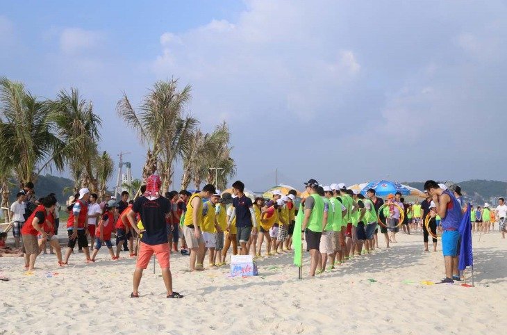 Một nhóm người tham gia trò chơi teambuilding trên bãi biển với cát trắng, cây dừa, bầu trời nắng đẹp và khán giả xung quanh.