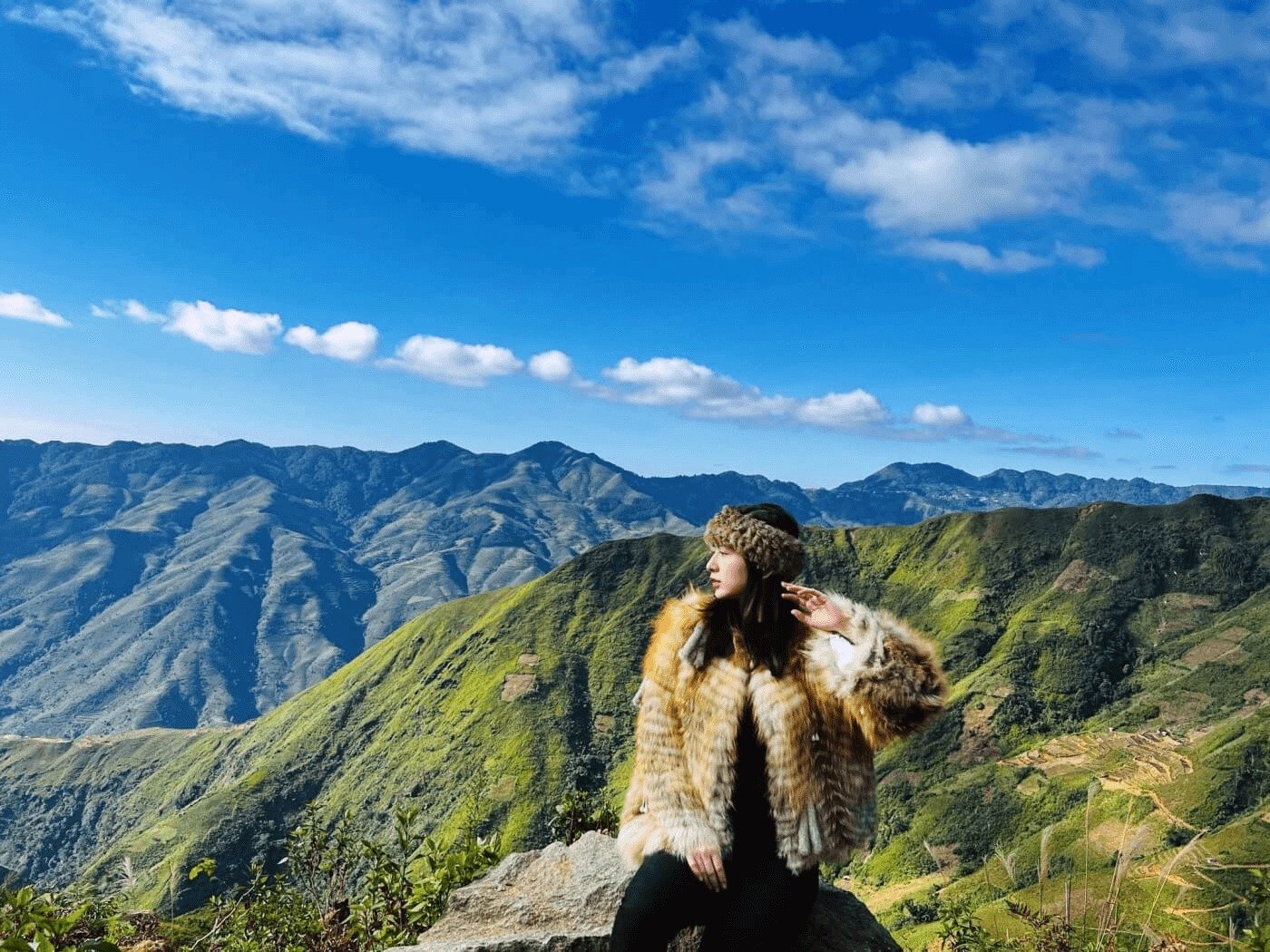 Người phụ nữ đeo mũ và áo lông đứng trên đồi xanh, ngắm cảnh núi non trùng điệp dưới bầu trời xanh.