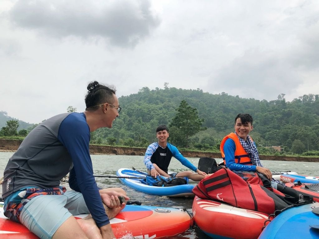 Ba người đàn ông vui cười ngồi trên ván chèo, mặc áo tắm, tận hưởng chuyến du lịch dưới trời mây cùng khung cảnh thiên nhiên xanh tươi.