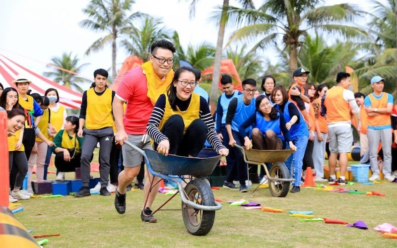 Nhóm đông người đang tham gia hoạt động teambuilding ngoài trời, chơi trò đua xe cút kít với không khí vui vẻ, đầy hào hứng.