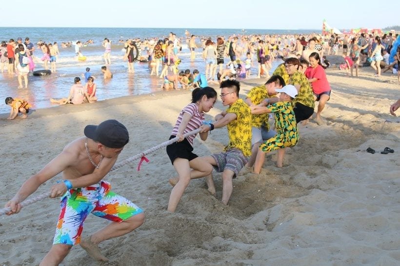 Một nhóm người tham gia trò kéo co trên bãi biển đông đúc, dưới ánh nắng và gần mặt nước.