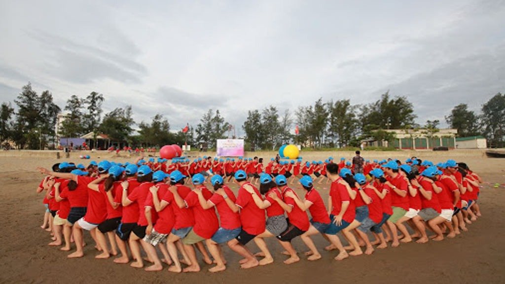 Nhóm người mặc áo đỏ và đội mũ xanh tham gia hoạt động tập thể trên bãi biển, với cát, cây cối và trời mây phía sau.