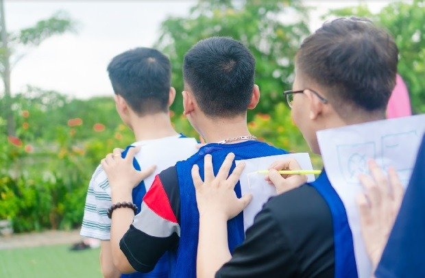 Nhóm người tham gia hoạt động ngoài trời, xếp hàng và vẽ lên giấy dán trên lưng người phía trước, khung cảnh thiên nhiên xanh mát.