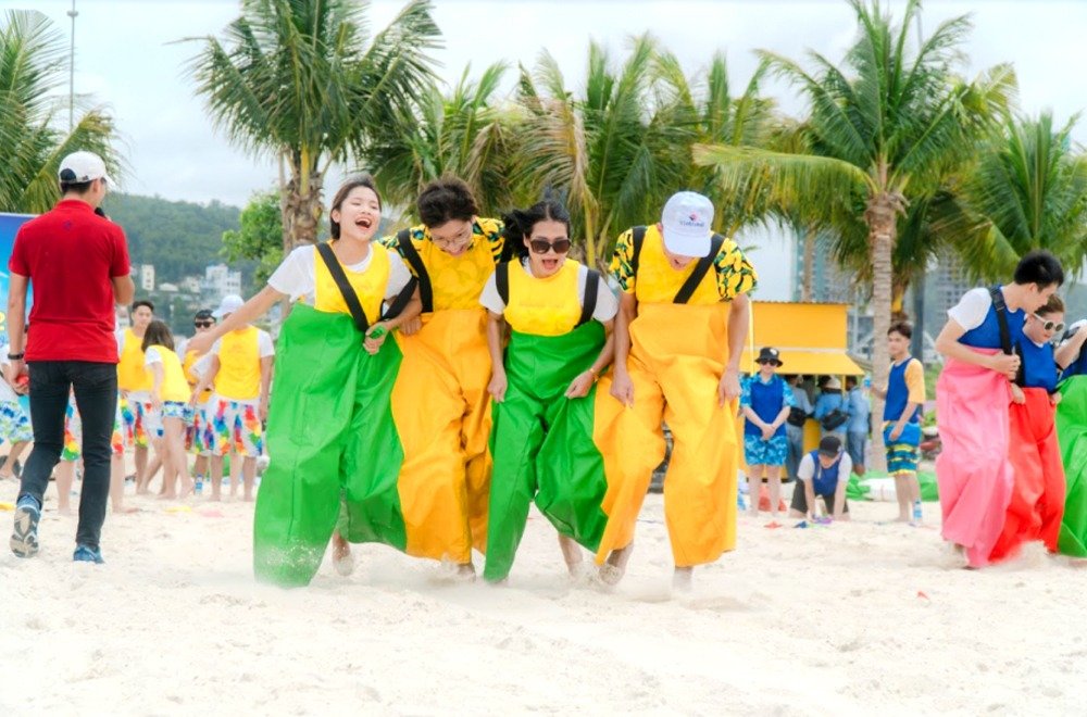 Nhóm bạn trẻ tham gia trò chơi đua bao trên bãi biển, tạo không khí vui nhộn giữa khung cảnh nhiệt đới xanh mát.