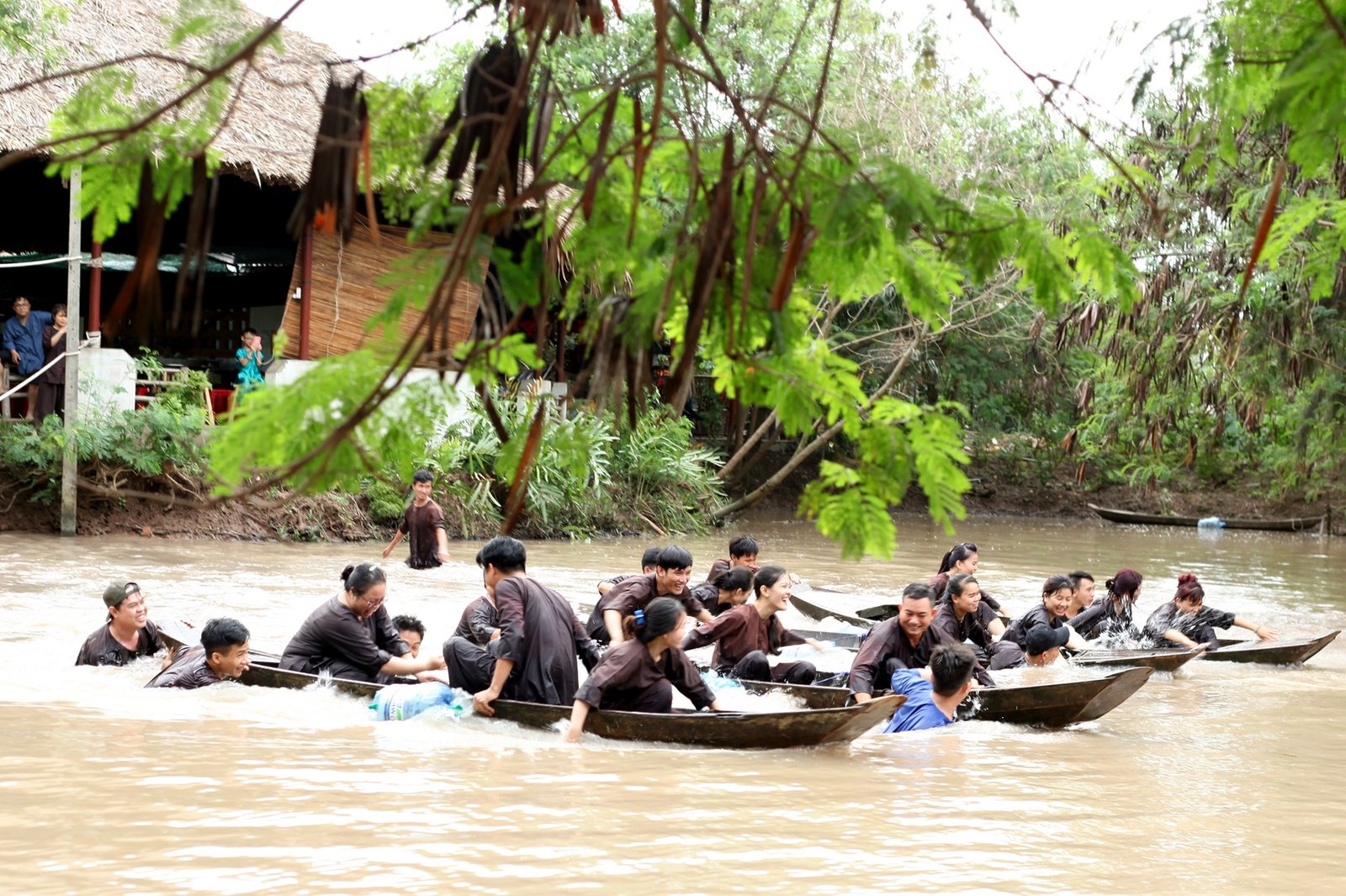 Người dân vui chơi trên sông bằng thuyền nhỏ ở miền quê Việt Nam, bên cạnh nhà sàn và cây xanh.