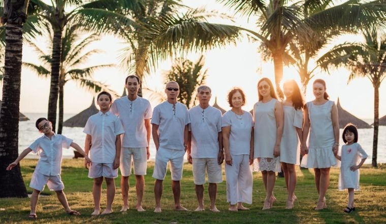 Gia đình lớn chụp ảnh kỷ niệm bên bờ biển lúc hoàng hôn, tất cả đều mặc đồ trắng dưới những tán cây dừa xanh mát.