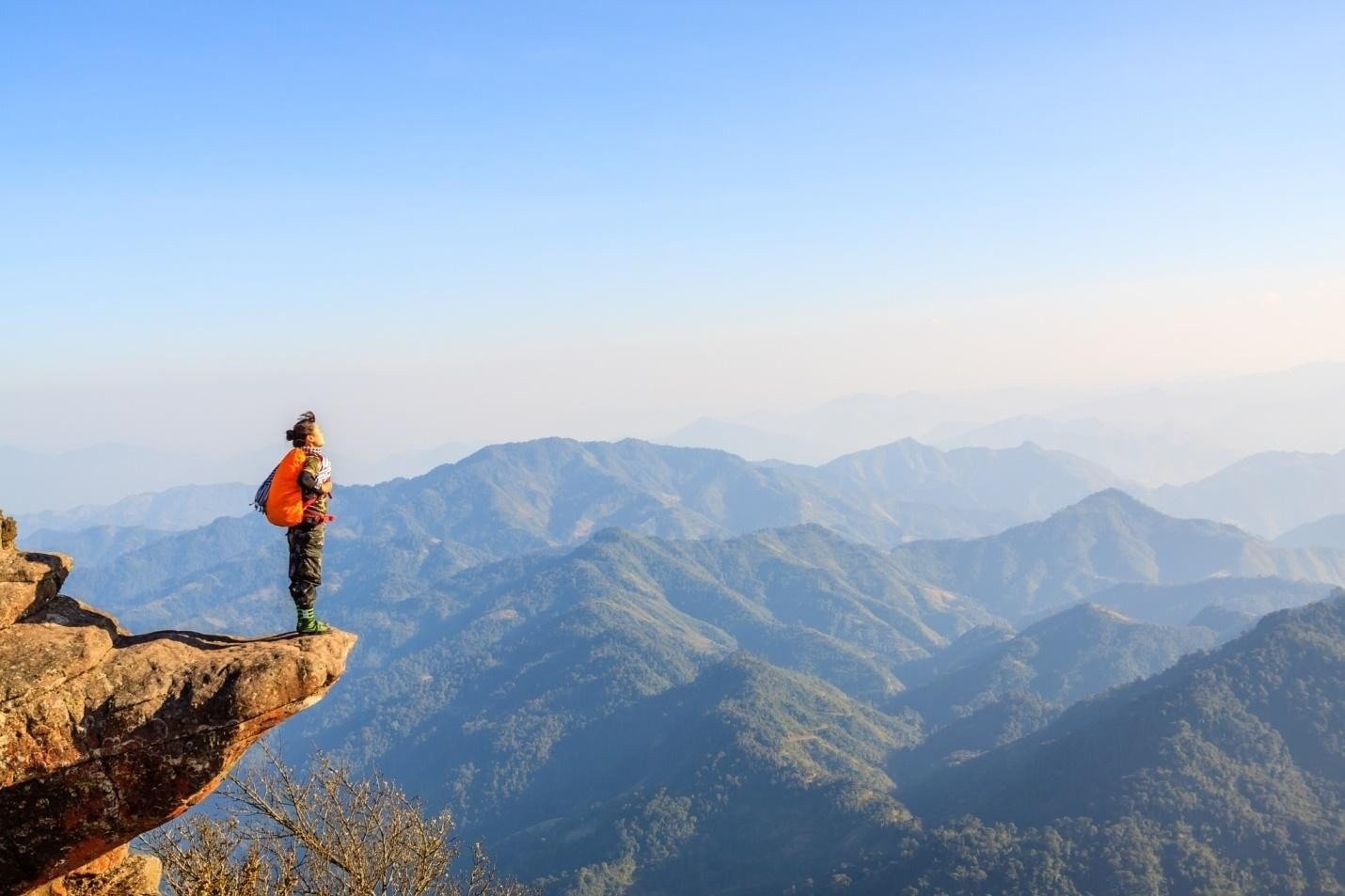 Người leo núi với balo cam đứng trên mỏm đá ngắm nhìn phong cảnh núi non hùng vĩ dưới bầu trời trong xanh.