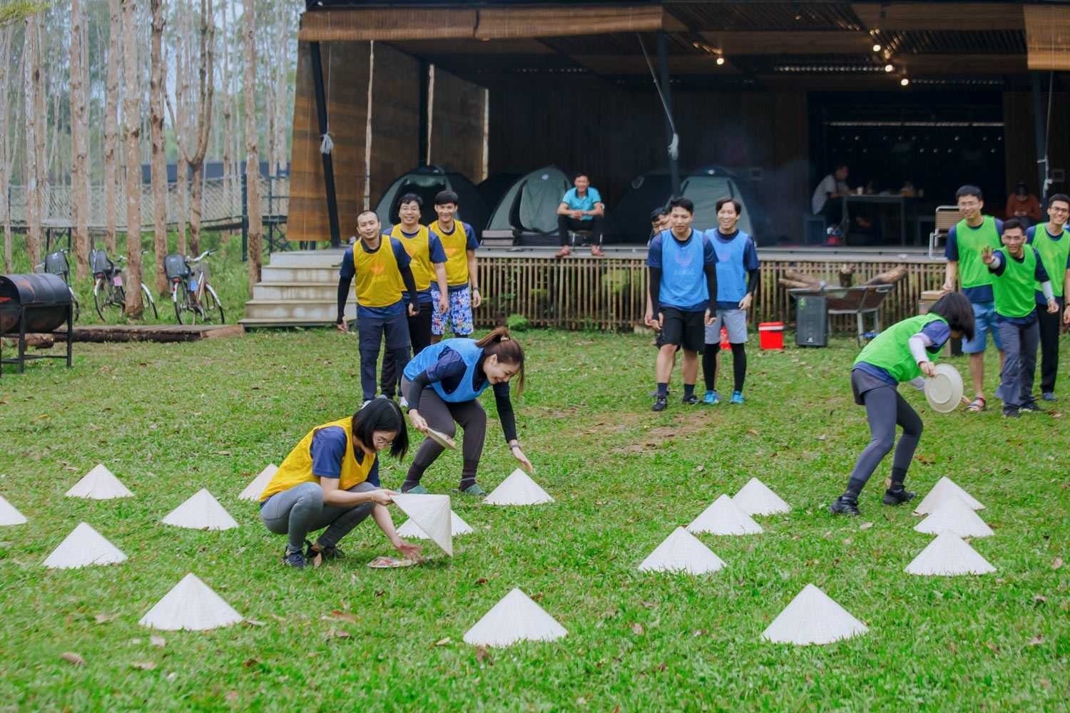 10 Lợi Ích Bất Ngờ Của Team Building Mà Bạn Chưa Biết 5 Trò chơi tập thể ngoài trời với người chơi đeo áo bib vàng, xanh lật nón lá, sân cỏ, xe đạp, lều trại và cây cối.