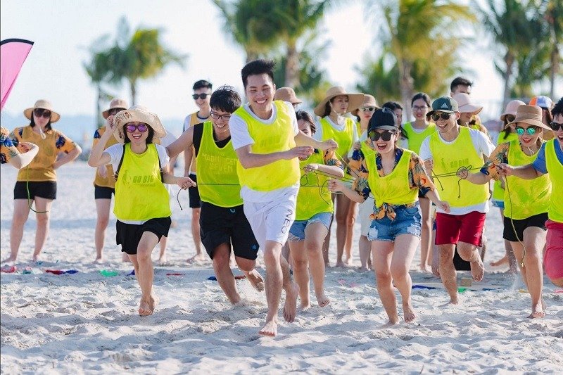 10 Lợi Ích Bất Ngờ Của Team Building Mà Bạn Chưa Biết 4 Một nhóm người vui vẻ tham gia trò chơi teambuilding trên bãi biển cát trắng, khung cảnh cây cọ xanh mát.