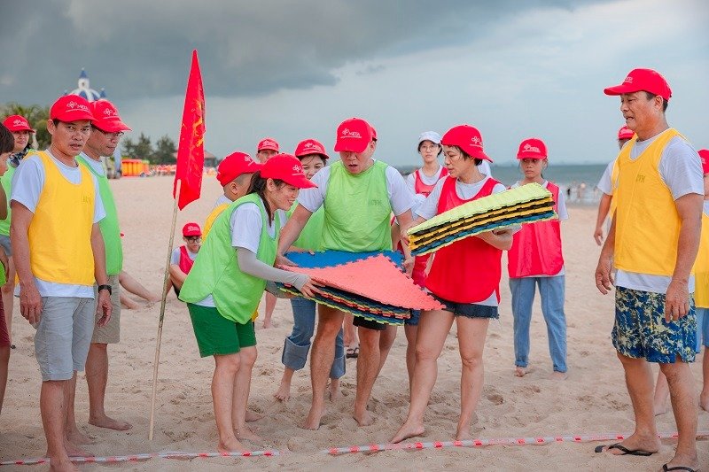10 Lợi Ích Bất Ngờ Của Team Building Mà Bạn Chưa Biết 2 Một nhóm người tham gia hoạt động team building trên bãi biển, đội mũ đỏ và mang áo phông màu sắc, cầm thảm trải.