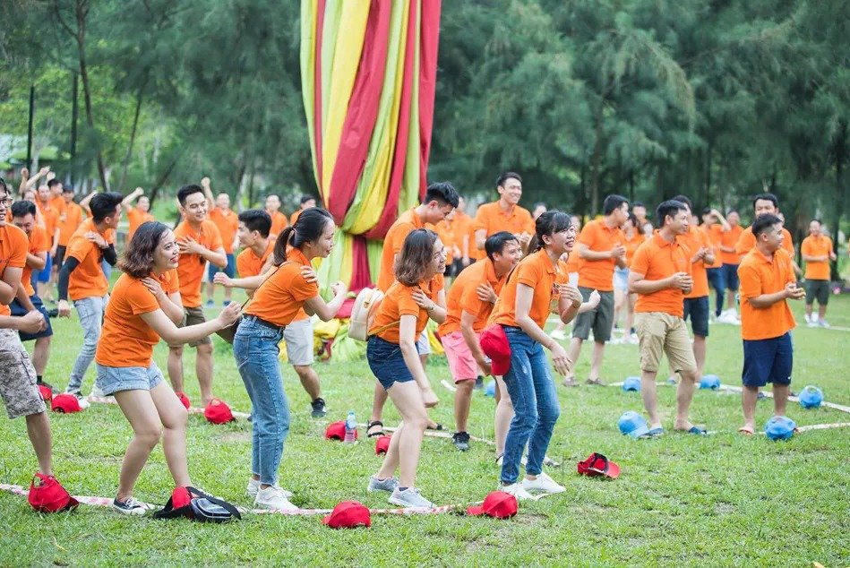 Nhân viên công ty mặc áo cam tham gia hoạt động nhóm ngoài trời, chơi trò chơi và cười đùa trong một buổi team building.