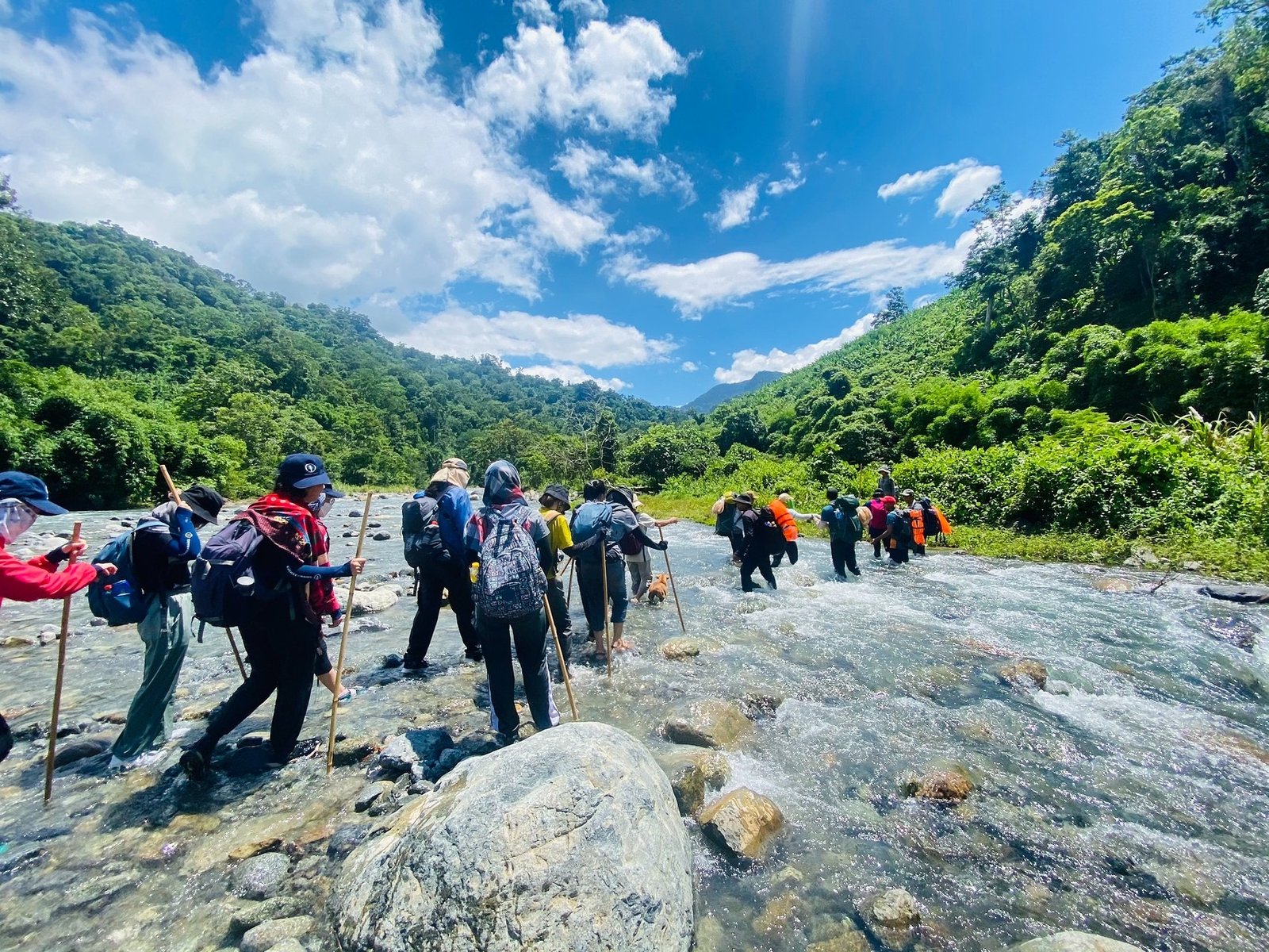 Một nhóm người đi bộ xuyên rừng, lội qua dòng suối trong một khung cảnh thiên nhiên oai hùng với bầu trời xanh ngát.