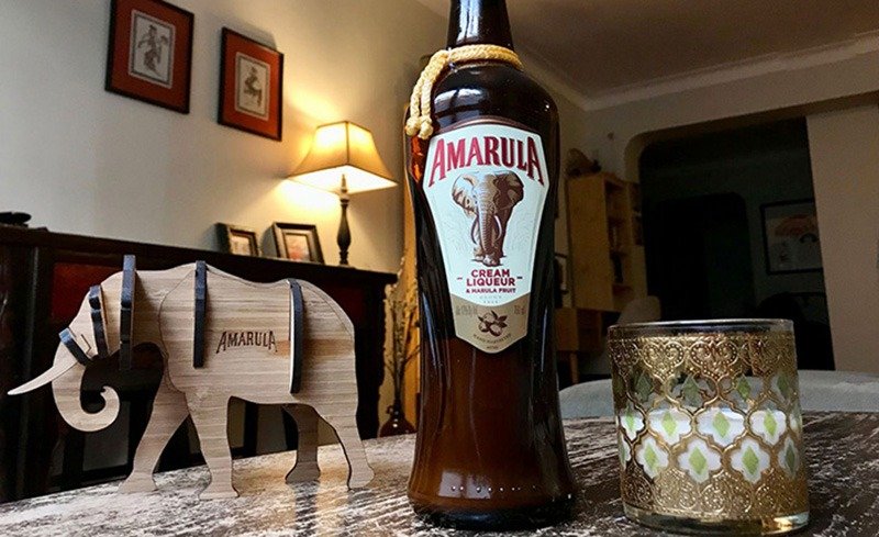 Chai Amarula, mô hình voi, ly trang trí vàng, trên bàn trong phòng khách ấm cúng được chiếu sáng bởi đèn.