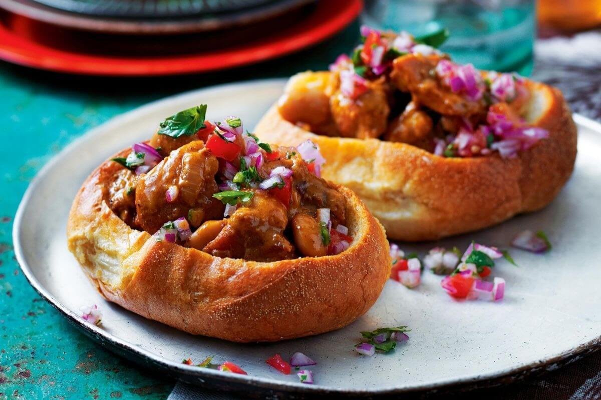 Review Nam Phi: Những Kinh Nghiệm Du Lịch Thú Vị 3 Bánh mì khoét rỗng chứa cà ri gà hấp dẫn, trang trí hành tím và rau thơm trên đĩa kim loại, bối cảnh sắc sảo.