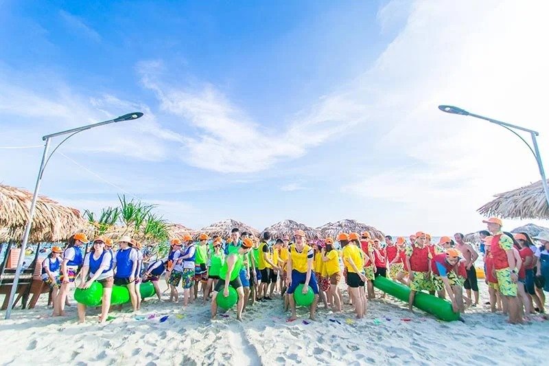 Nhóm người mặc áo khoác màu sắc trong hoạt động team building trên bãi biển, tạo không khí vui tươi dưới bầu trời xanh.