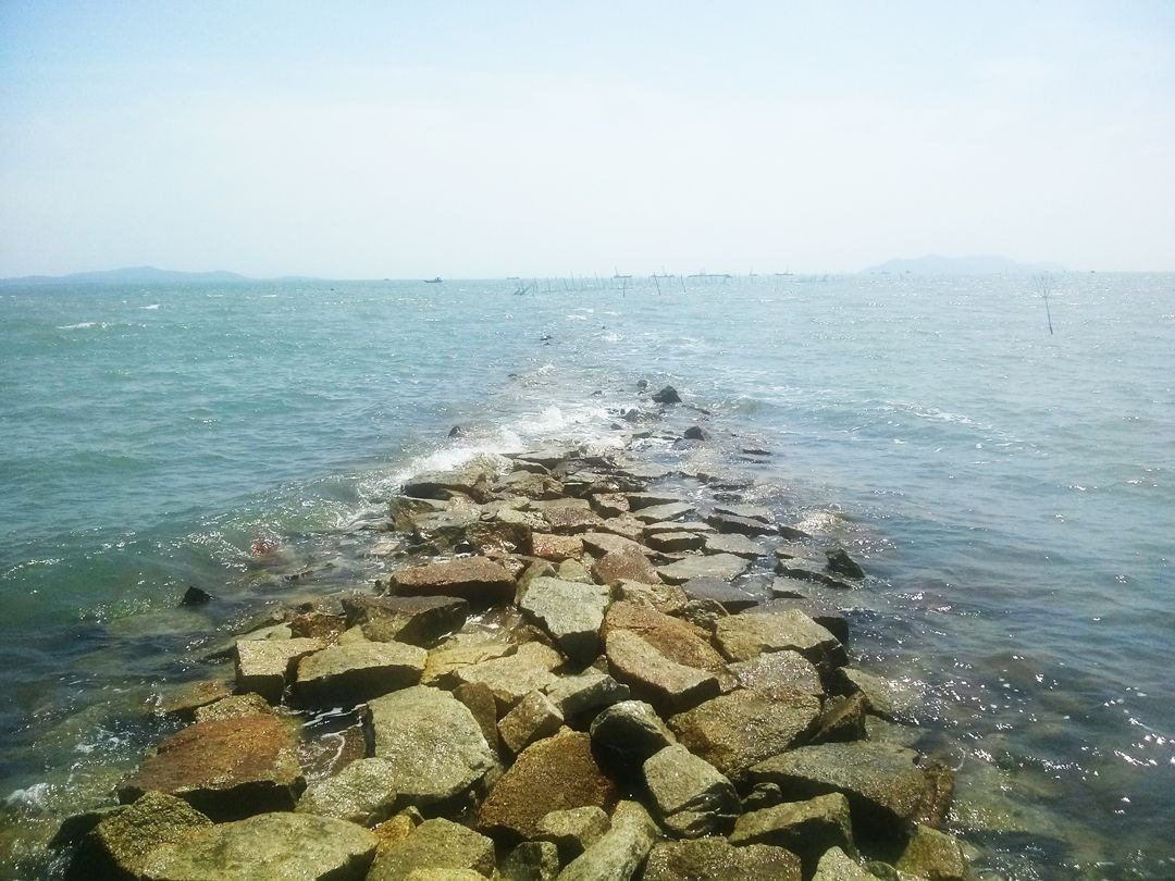 Hinh ảnh tường đá nhô ra biển xanh trong ngày nắng, sóng biển nhẹ nhàng đập vào đá tạo khung cảnh thiên nhiên thanh bình.