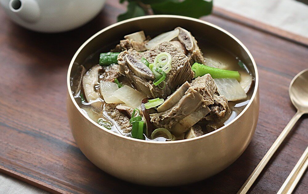Bát canh sườn bò Hàn Quốc với hành lá và củ cải, bày trên khay gỗ, kết hợp thìa và ấm trà trắng ở nền.