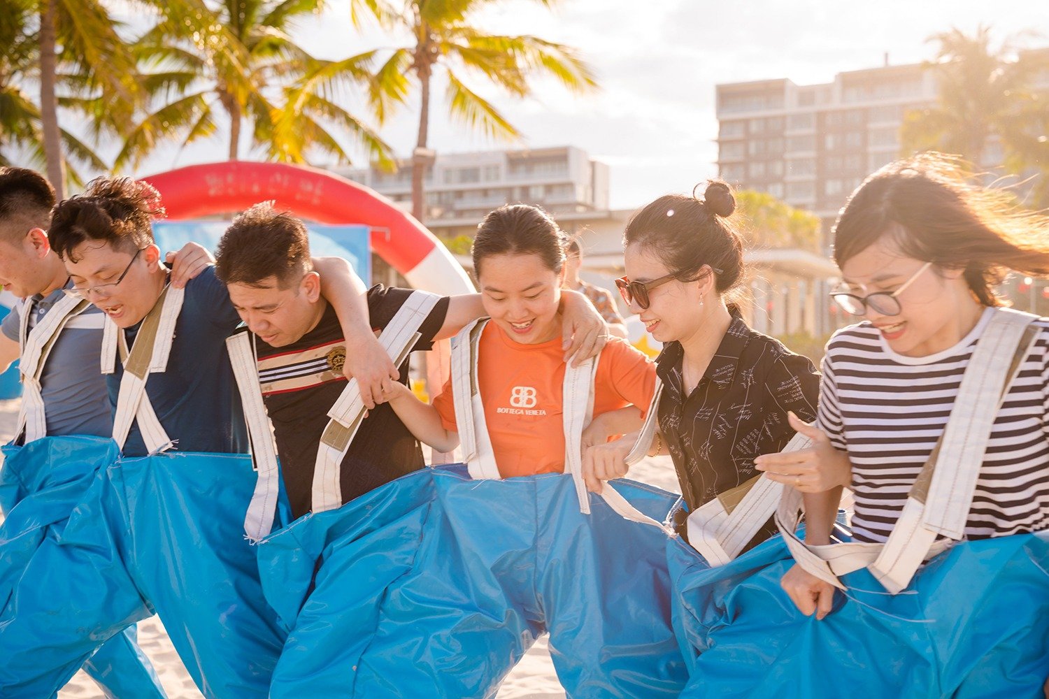 Nhóm người tham gia trò chơi team building trên bãi biển đầy nắng với cây dừa và tòa nhà phía xa.