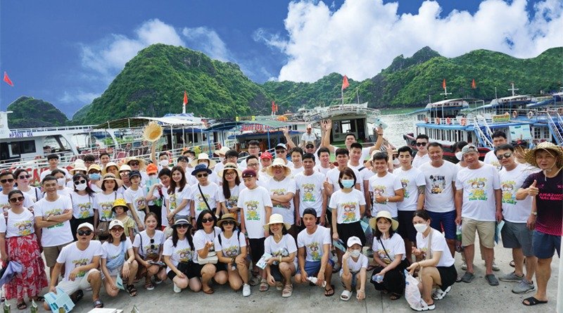 Nhóm người mặc áo trắng, đứng trước cảnh biển và núi xanh, có nhiều tàu đậu, bầu trời xanh thẳm và mây trắng.