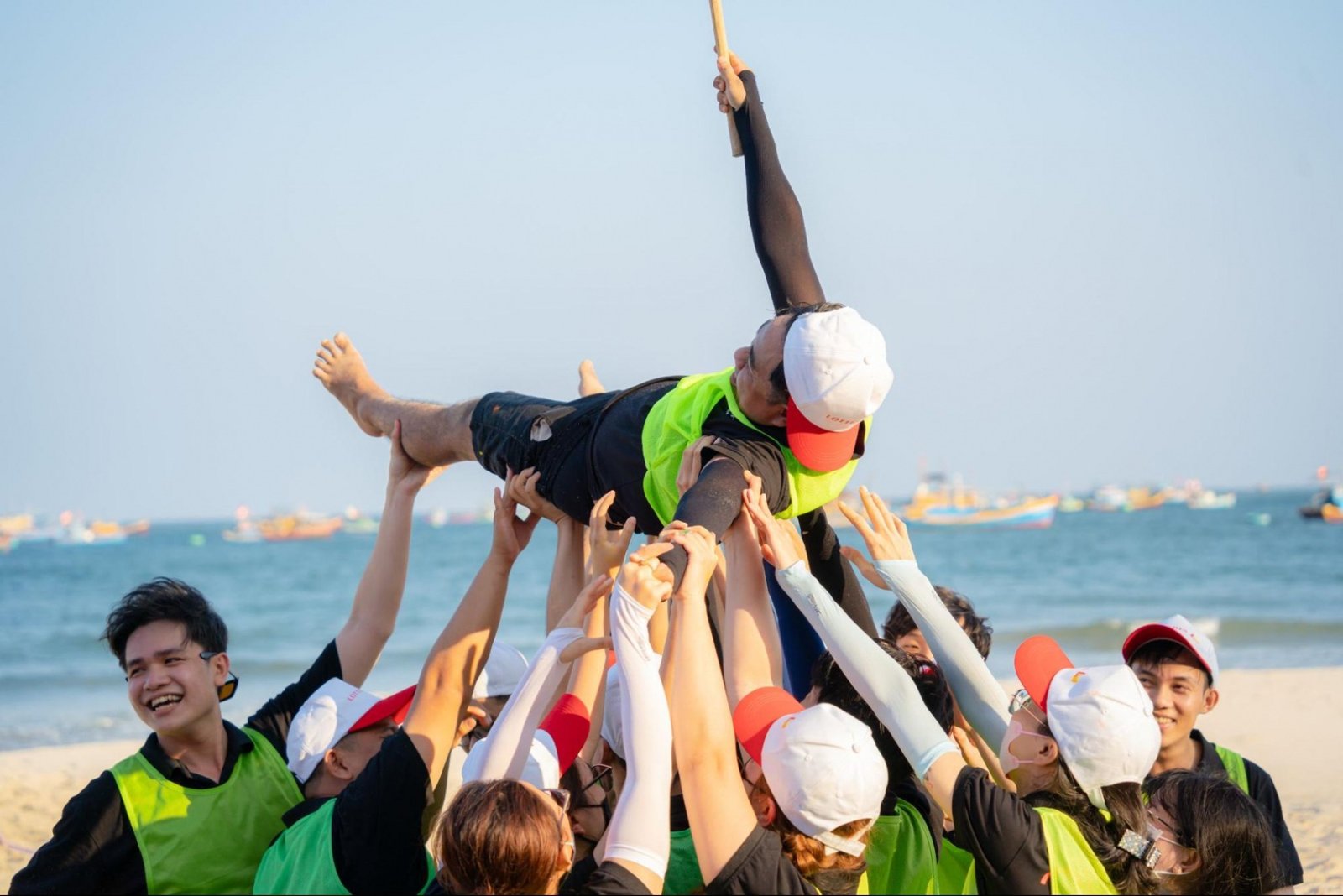 Nhóm người mặc áo xanh và nón trắng vui mừng nâng một người lên trong hoạt động teambuilding ngoài biển.