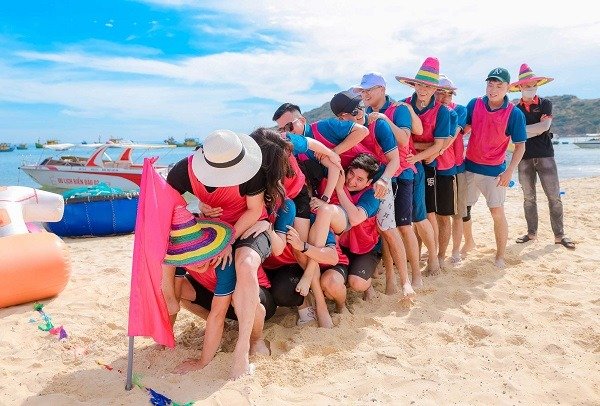 Nhóm người tham gia trò chơi teambuilding vui nhộn trên bãi biển, mặc áo thun đỏ và đội mũ lưỡi trai kèm mũ truyền thống.