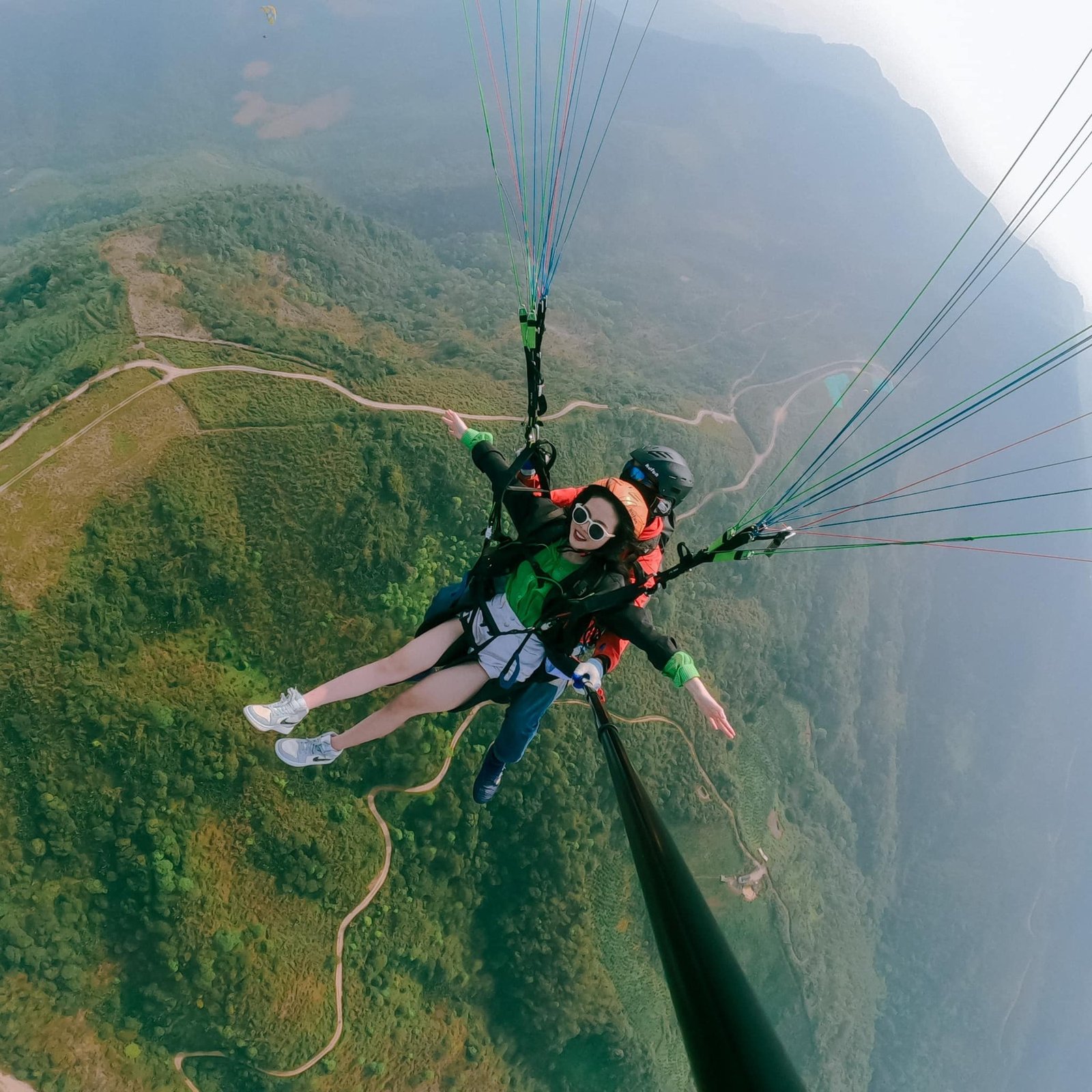 Người phụ nữ hào hứng trải nghiệm dù lượn đôi trên cao, khung cảnh núi non và thảm rừng xanh trải dài dưới chân.