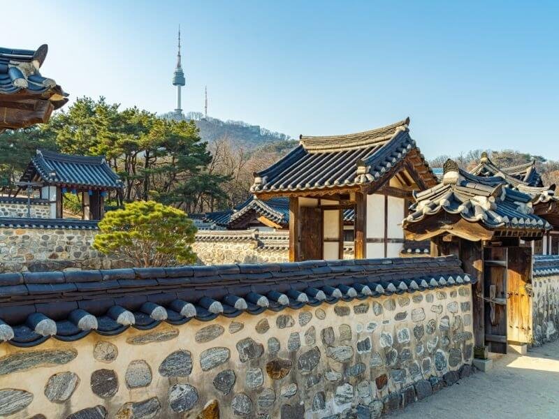 Ngôi nhà truyền thống Hàn Quốc với mái ngói đen, tường đá, cây xanh và tháp truyền hình Namsan ở hậu cảnh.