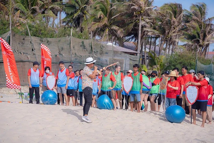 Nhóm người chơi trò chơi team building trên bãi biển, cây cối xung quanh, không khí vui vẻ và sôi động.