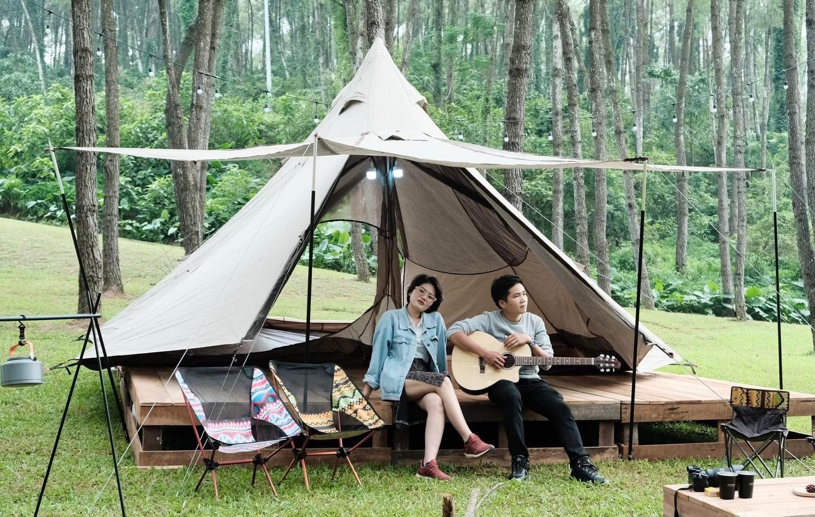 Cặp đôi ngồi trước lều cắm trại giữa rừng thông xanh, người đàn ông chơi guitar trong không gian thiên nhiên thư giãn.