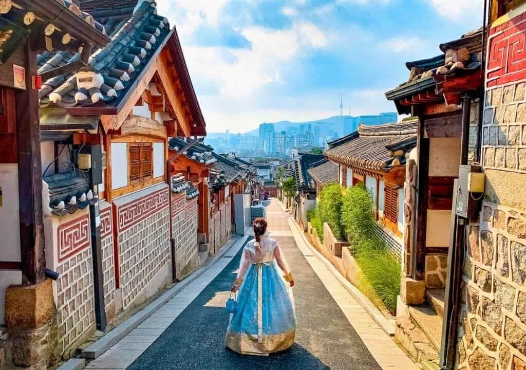 Người mặc hanbok dạo bước trên con đường truyền thống Bukchon Hanok, Seoul, với cảnh quan thành phố phía xa.
