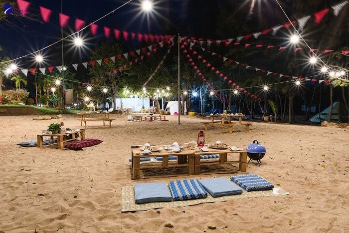 Bãi biển về đêm với ánh đèn lung linh, bàn ghế gỗ và đệm ngồi, tạo không gian thư giãn và lãng mạn.