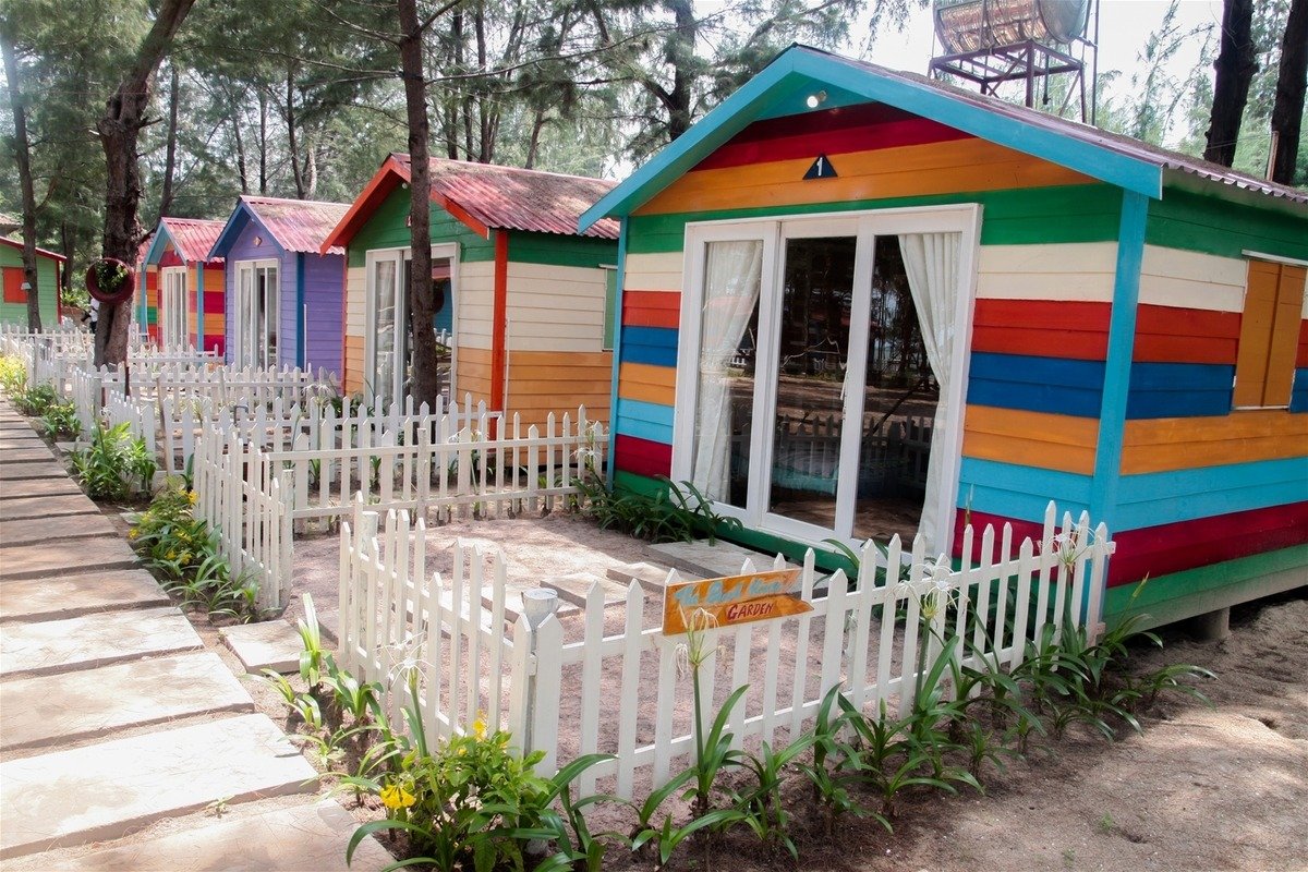 Dãy nhà nhỏ nhiều màu sắc bên hàng rào trắng, nằm trong rừng cây xanh mát, tạo cảm giác vui tươi và yên bình.