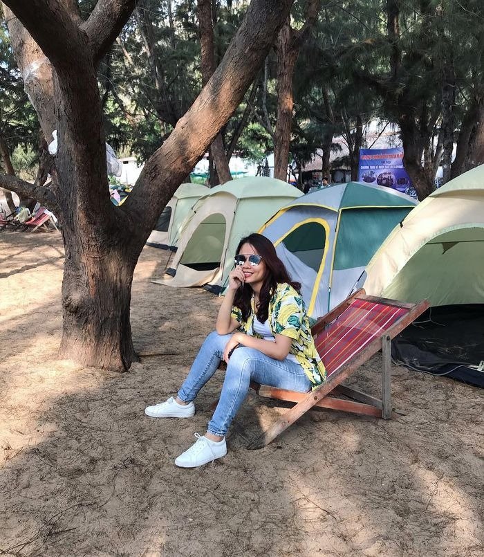 Người phụ nữ ngồi thư giãn trên ghế dựa tại khu cắm trại, xung quanh là lều xanh và cây cổ thụ che bóng mát.