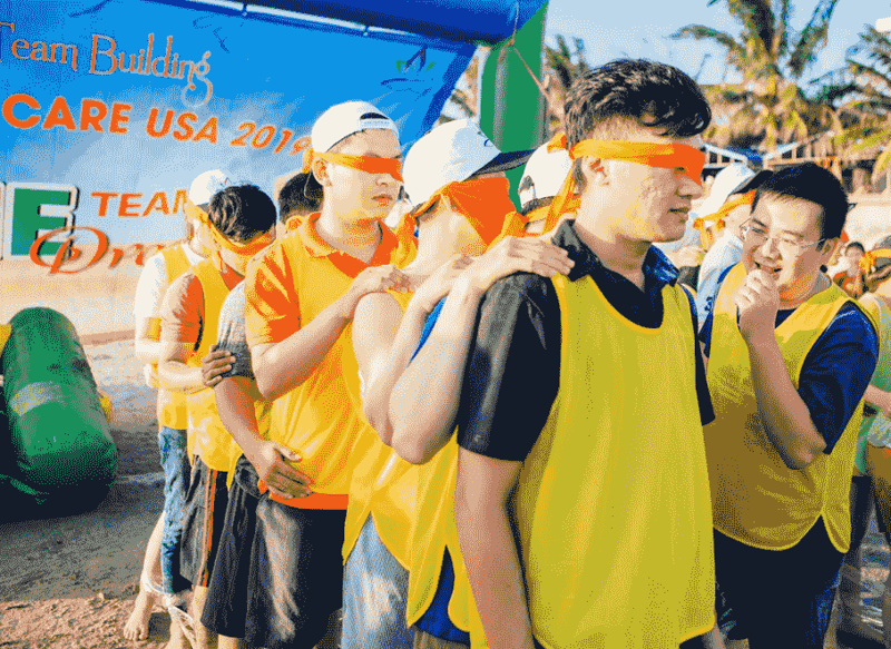 Nhóm người tham gia hoạt động team building, bịt mắt và mặc áo thun vàng, tại sự kiện ngoài trời năm 2019.