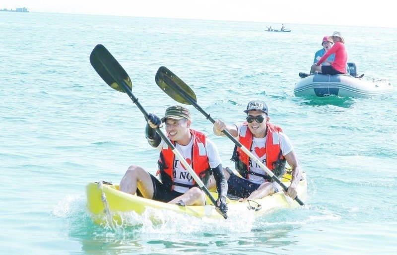 Hai người chèo thuyền kayak mặc áo phao cam tươi cười, chèo trên biển xanh trong ánh sáng mặt trời rực rỡ.