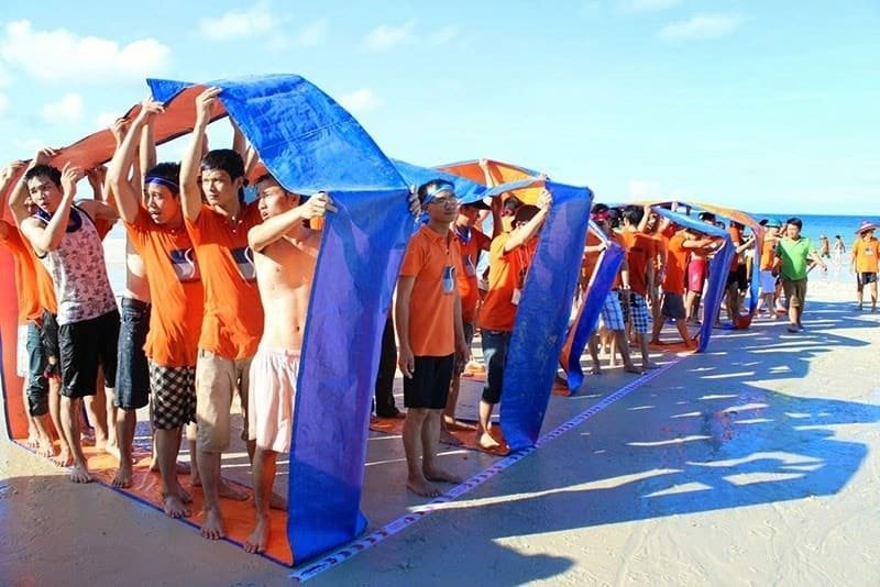 Nhóm người tham gia trò chơi team building trên bãi biển với áo cam, băng đầu xanh và thảm màu xanh da trời.