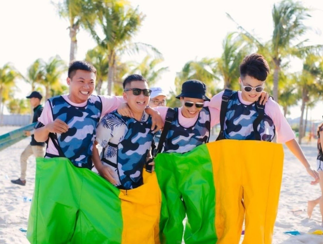 Nhóm bạn trẻ đang tham gia trò chơi trên bãi biển dưới ánh nắng, mặc áo họa tiết rằn ri và quần xanh vàng.