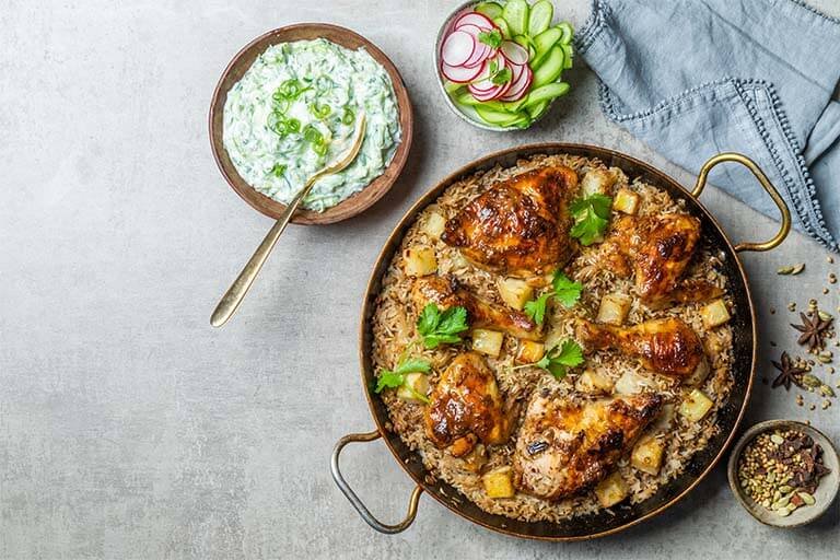 Món ăn gồm gà nướng, cơm, rau mùi và khoai tây, kèm sốt kem và salad dưa chuột, củ cải thái lát trên nền xám.