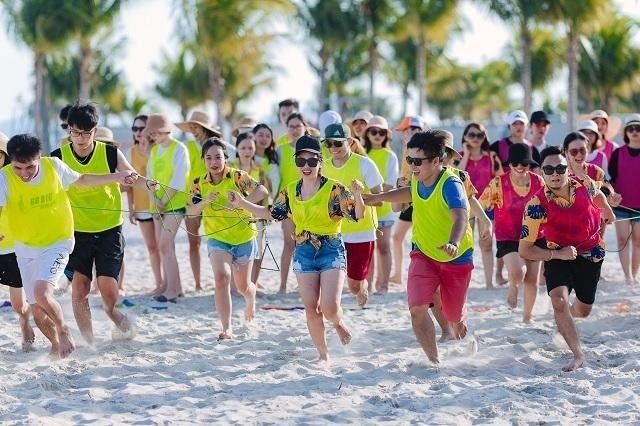 Nhóm người mặc áo thun neon vui chơi và chạy trên bãi biển đầy nắng, có nhiều cây cọ phía sau.