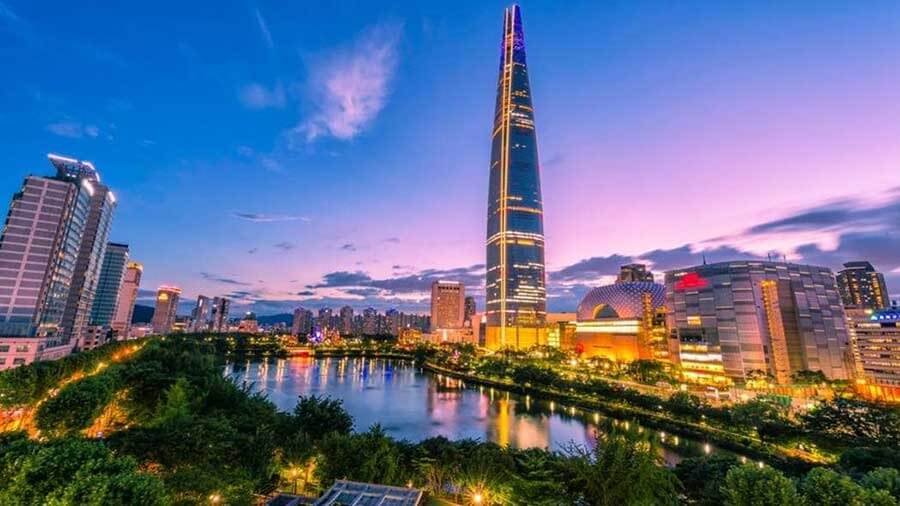 Tháp cao chọc trời bên hồ nước phản chiếu ánh đèn rực rỡ khi hoàng hôn, với bầu trời xanh nhạt và mây nhẹ nhàng.