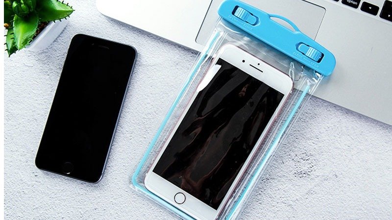 Top 5 Kinh Nghiệm Du Lịch Biển Cho Chuyến Đi Hoàn Hảo 5 Điện thoại nằm trong túi chống nước màu xanh, kế bên là điện thoại khác trên bàn laptop và cây cảnh nhỏ.